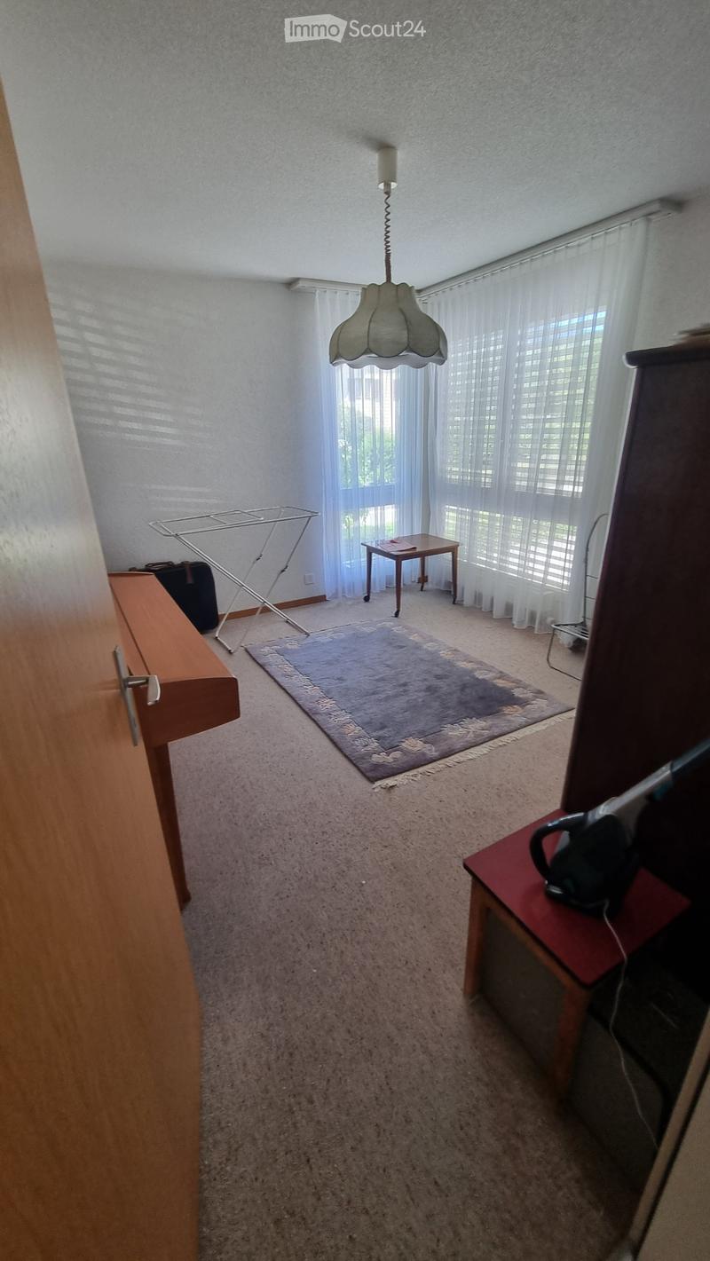 A small, carpeted room with a wooden door, a small table with a chair, an ironing board, and a hanging lamp.