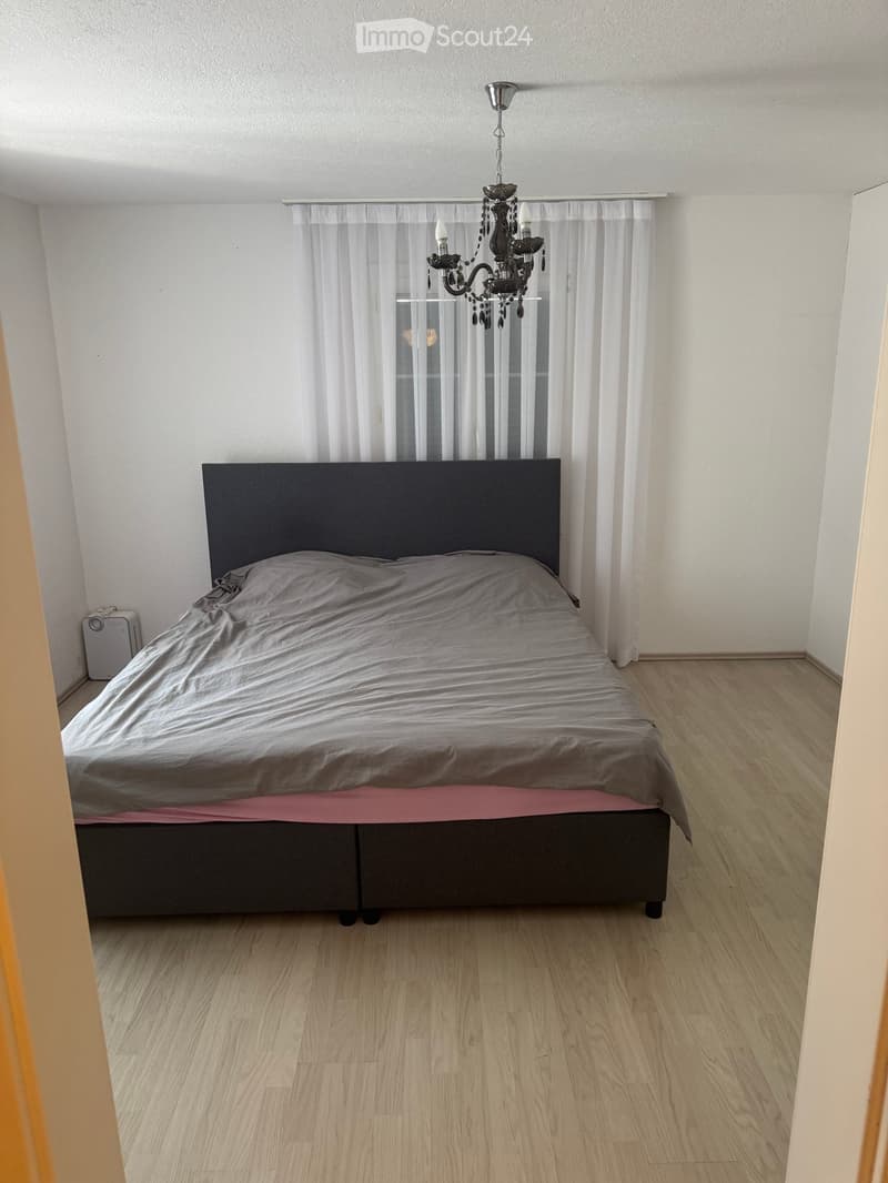 A modern bedroom with a wooden floor, a large bed with grey bedding, a dark headboard, and a chandelier hanging from the ceiling. White curtains cover the window.
