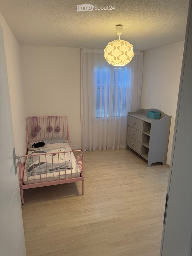 Cozy bedroom with a pink metal bed, grey nightstand, and a modern hanging lamp. The room features wooden flooring and large windows with sheer curtains.