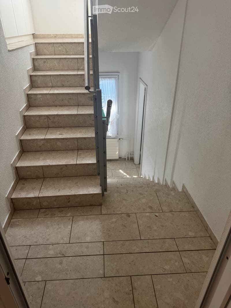 Staircase with beige tiled steps and walls, metal handrail, and a window with white curtains at the top landing.