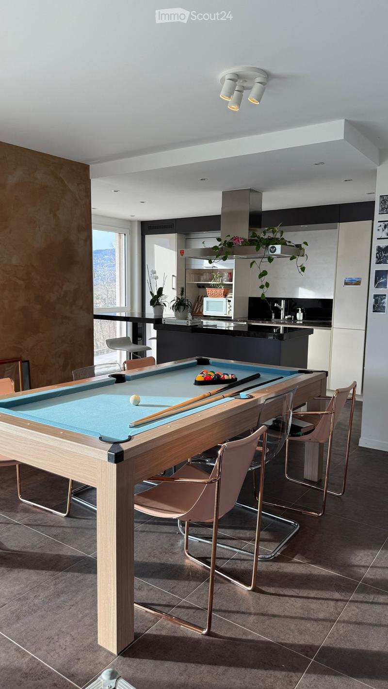 Modern kitchen with a pool table, bar stools, and a kitchen island. The space features a sleek design with a large window offering a scenic view.