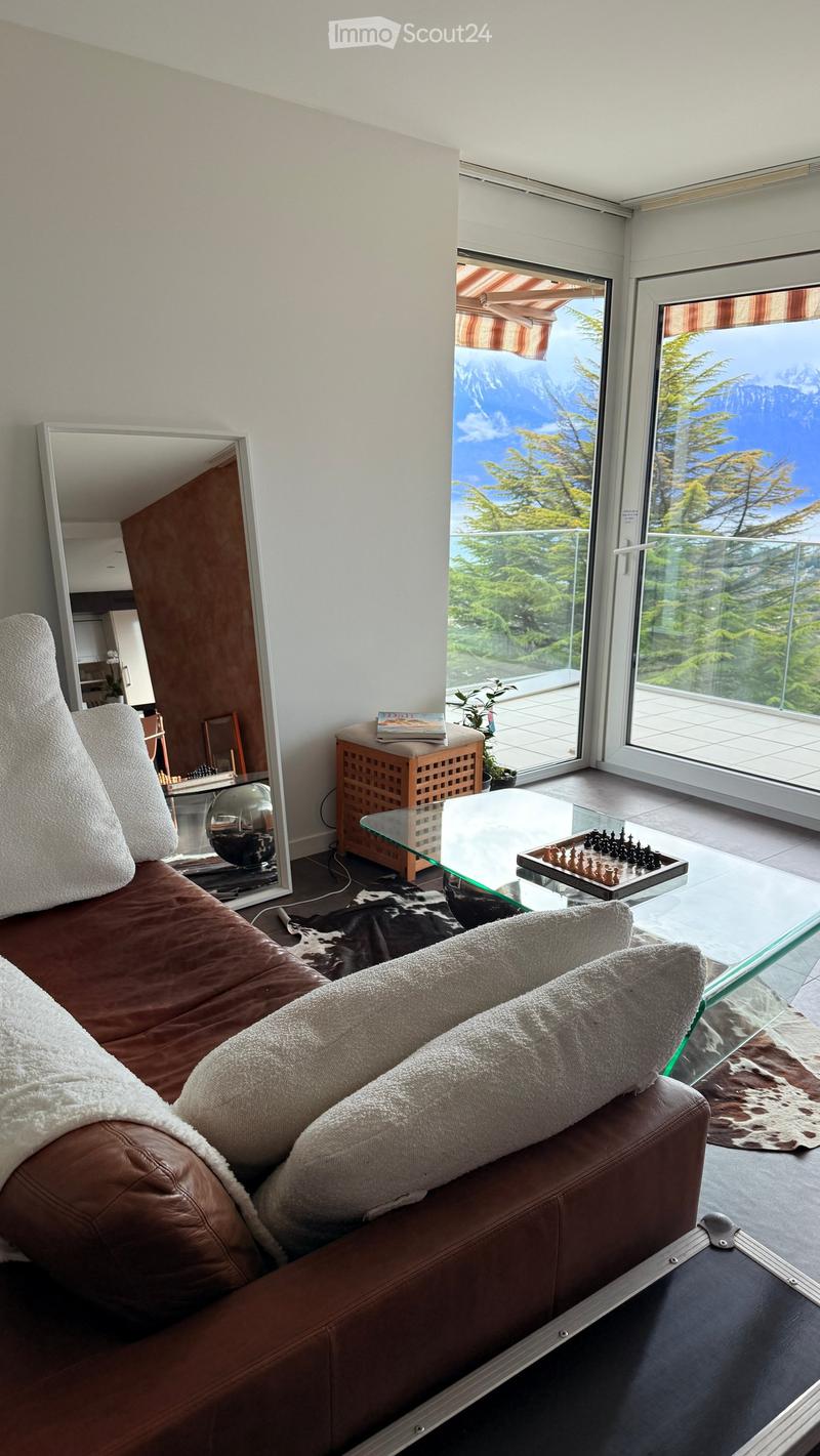 Modern living room with a brown leather sofa, glass coffee table, chessboard, large mirror, side table, and a scenic mountain view through large windows.