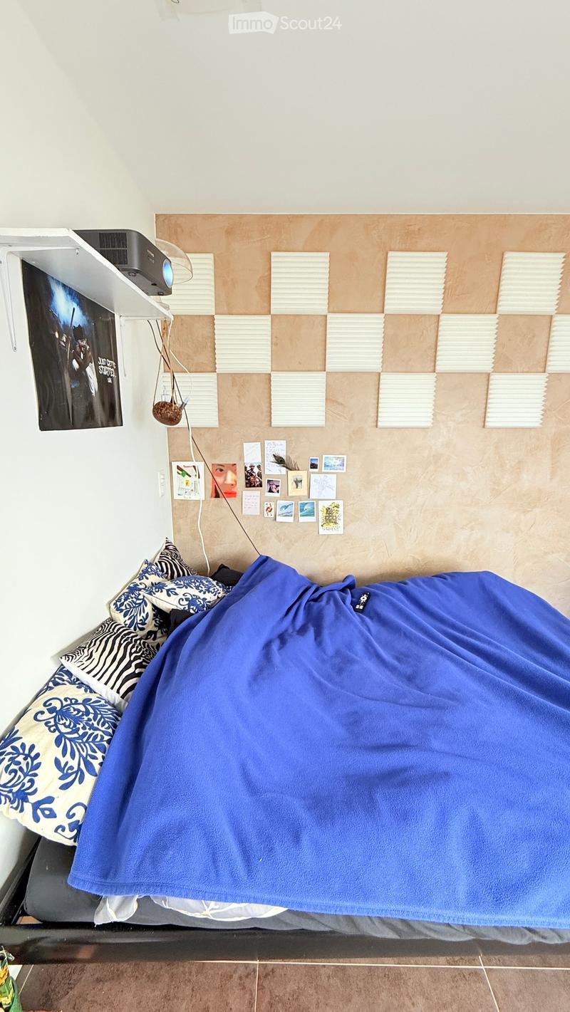 A cozy bedroom with a bed covered in a blue blanket and patterned pillows, a projector mounted on a shelf, and several picture frames on the wall.