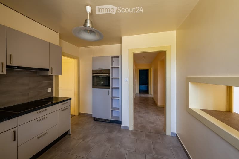 Empty hallway with tiled floor, light-colored walls, and a modern pendant light fixture. The hallway leads to a kitchen area with cabinets, a stove, and a refrigerator, and a small room visible at the end.