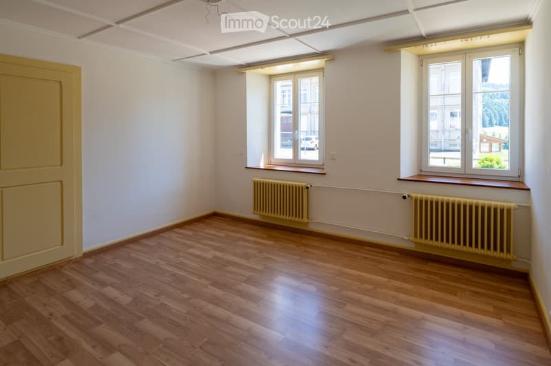 Empty room with wooden flooring, white walls, and two radiators. Features large windows with blinds and a door on the left side.