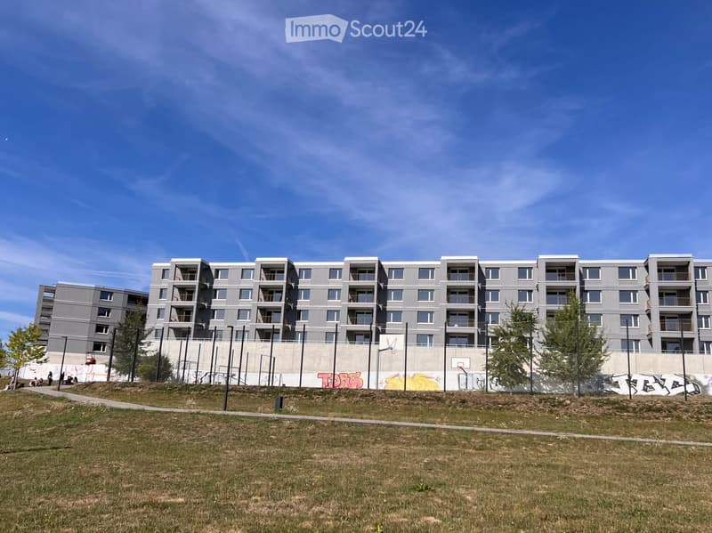 Modern multi-story apartment building with grey facade, multiple windows, and balconies, surrounded by a grassy area and a fence with graffiti.