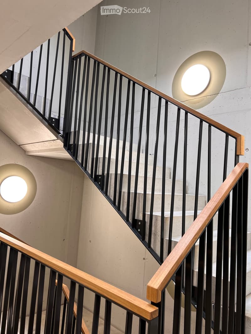 Modern staircase with wooden handrails and black metal balusters, illuminated by circular wall-mounted lights.
