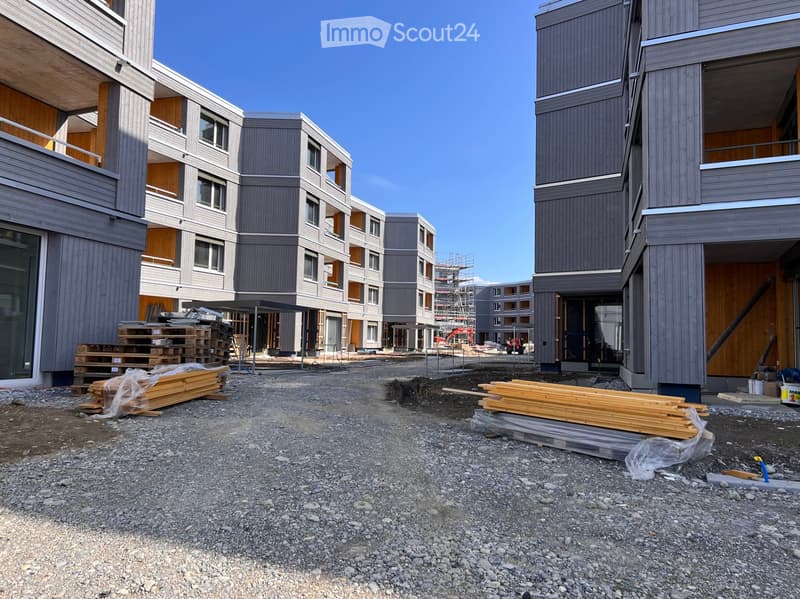 Modern multi-story residential building under construction with grey and wooden exterior, visible windows, and construction materials scattered around.