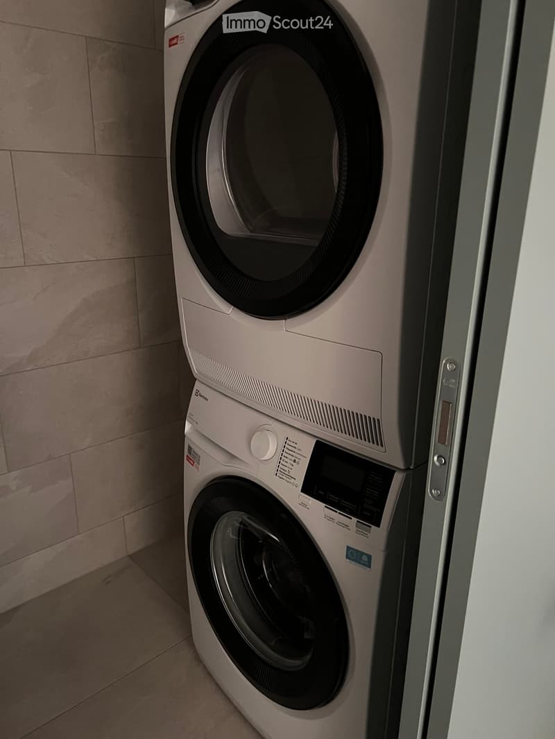 A modern, compact bathroom featuring a stacked washing machine and dryer unit with a sleek, white finish and digital control panel.