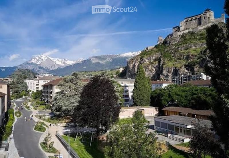 Panoramic view of a town with snow-capped mountains in the background, featuring residential buildings, trees, and a clear blue sky.