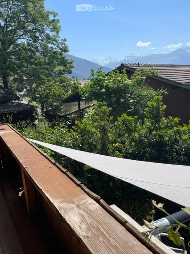 Balcony with wooden railing, overlooking a lush green garden and distant mountains under a clear blue sky.