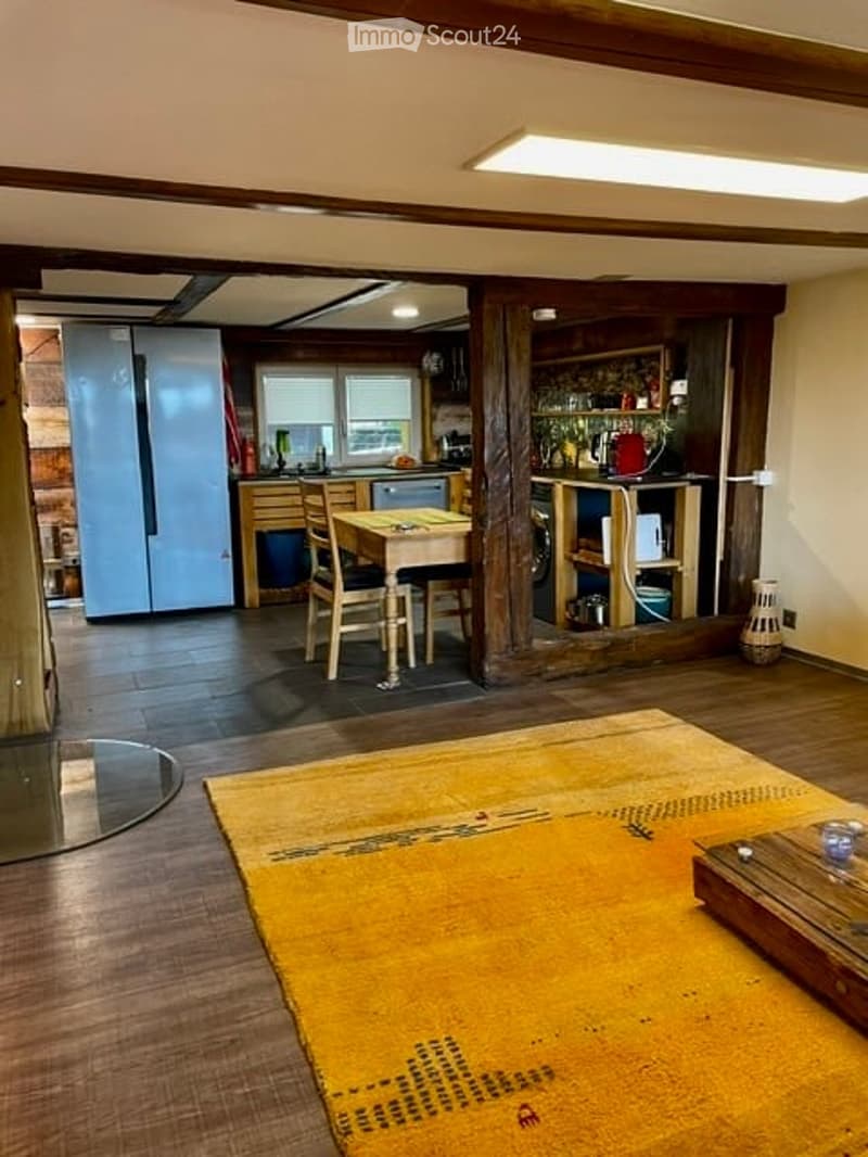 Open-plan living and dining area with a large yellow rug, wooden flooring, a sofa, coffee table, dining table with chairs, a bar area with bar stools and shelves, and a kitchen area with cabinets, counter, sink, and appliances.