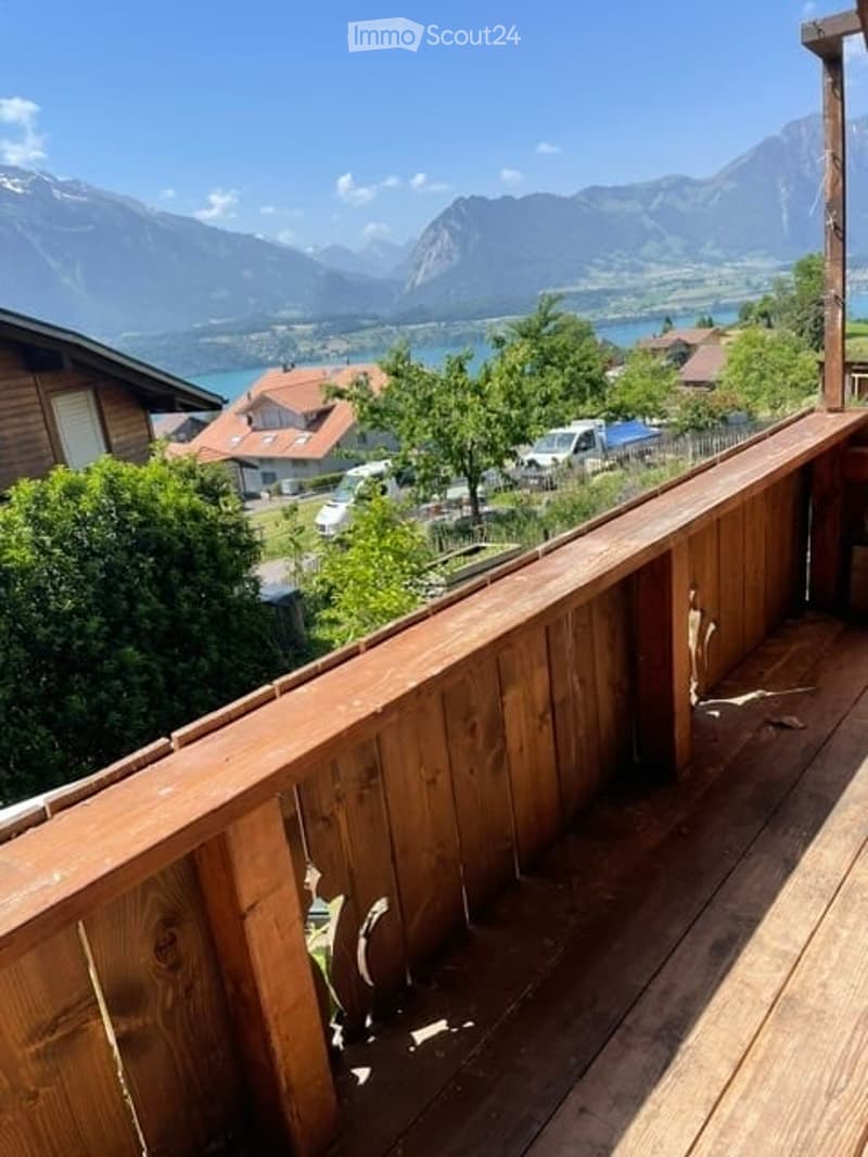 Balcony with wooden railing, overlooking a scenic mountain and lake view, surrounded by greenery and neighboring houses.