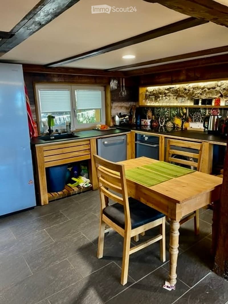 Cozy kitchen with wooden cabinets, a dining table with chairs, a refrigerator, dishwasher, and various kitchen utensils. The space features exposed wooden beams and a tiled floor.
