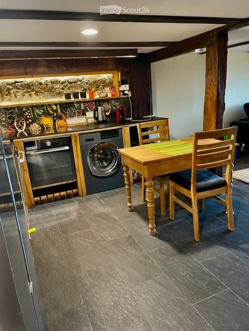 Modern kitchen with wooden beams, stone backsplash, built-in oven, washing machine, and a small dining area with a wooden table and chairs.