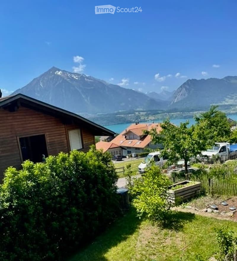 Scenic outdoor view of a wooden house surrounded by lush greenery, with a clear view of a mountain range and a lake in the background. Several parked vehicles and a small garden area are visible.