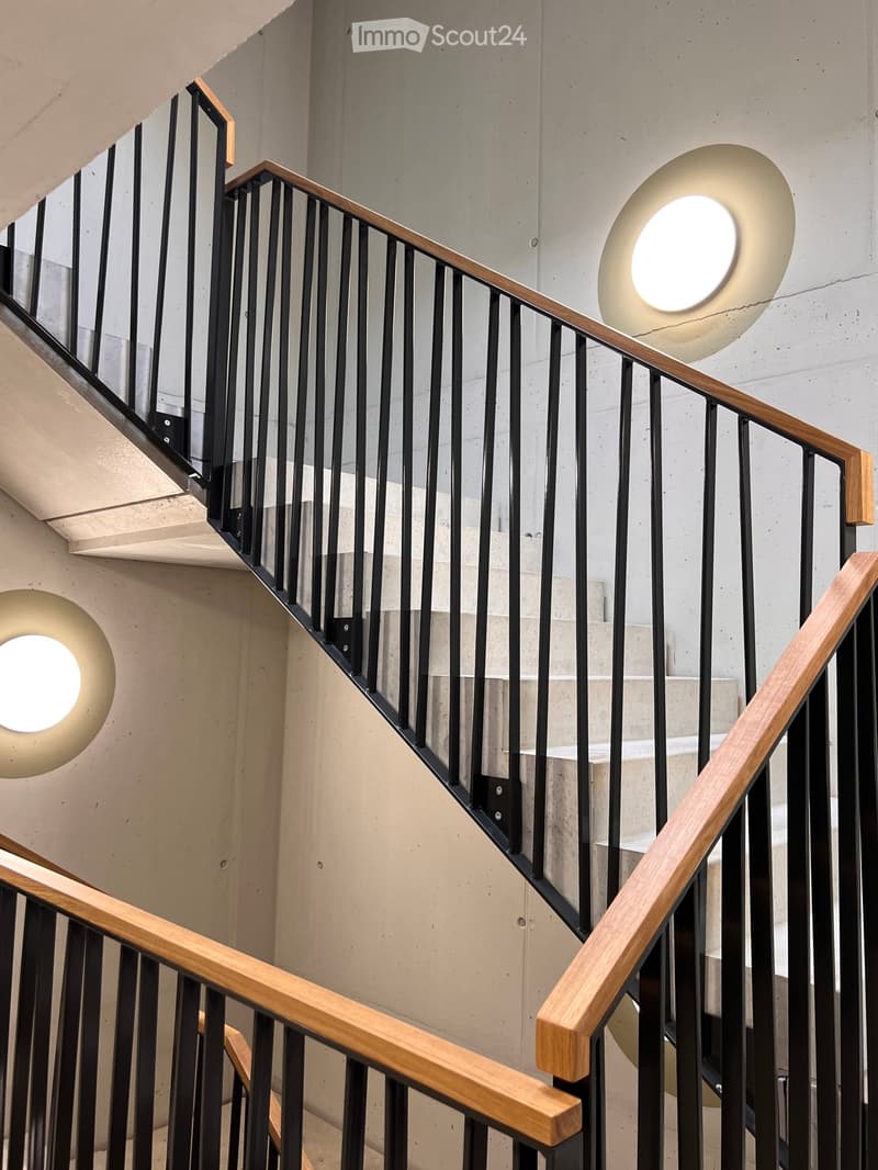 Modern interior staircase with wooden handrails and black metal balustrades, illuminated by circular wall-mounted lights.