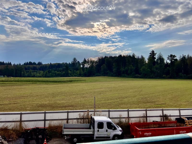 A wide, open field with a wooden fence in the foreground, a white van parked near the fence, and a red dumpster to the right. The background features a line of trees and a partly cloudy sky.