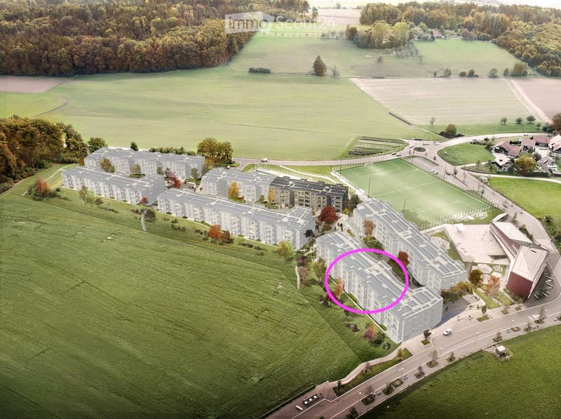 Aerial view of a residential complex with multiple apartment buildings, surrounded by green fields and trees. The buildings have solar panels on their roofs, and there is a soccer field and parking area nearby.