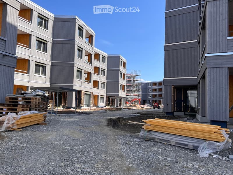 Modern multi-story residential building under construction with grey and brown exterior, visible windows, and construction materials scattered around.