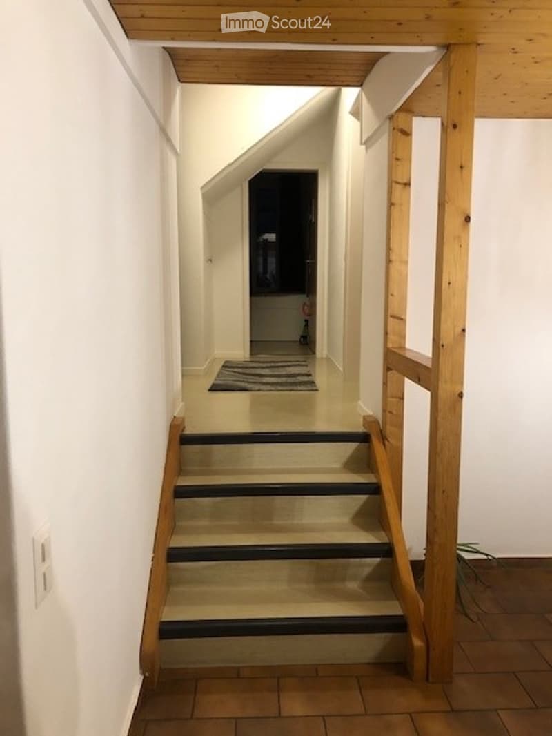 A narrow hallway with wooden stairs leading upwards, white walls, and a small rug at the base of the stairs.