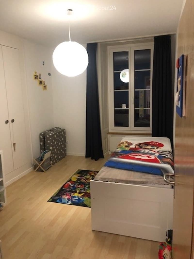 A small, well-lit bedroom with a single bed, colorful bedding, a wardrobe, a round hanging lamp, a colorful rug, and a suitcase near the window.