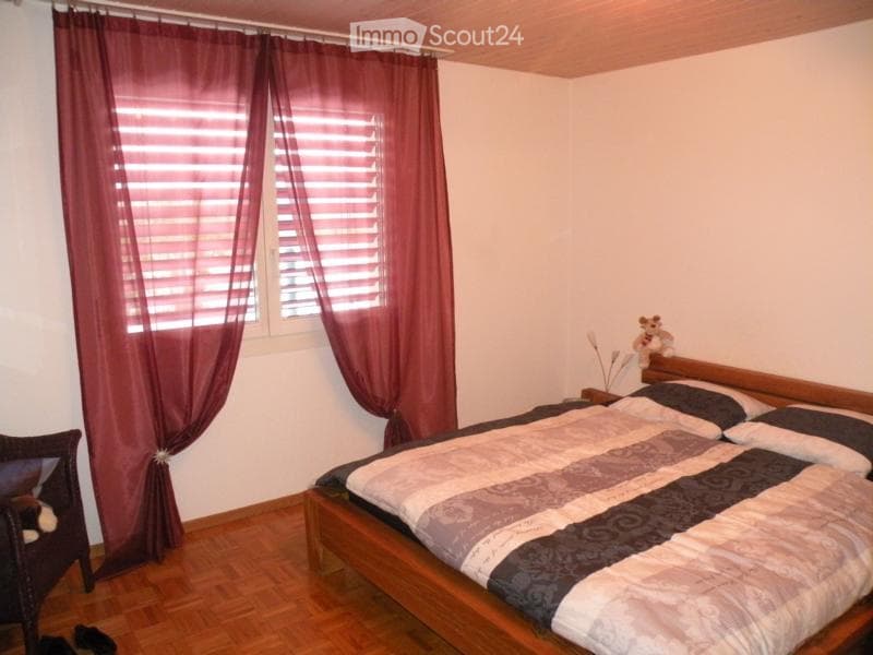 Cozy bedroom with wooden flooring, a large bed with patterned bedding, a wooden chair, and a window with red curtains.