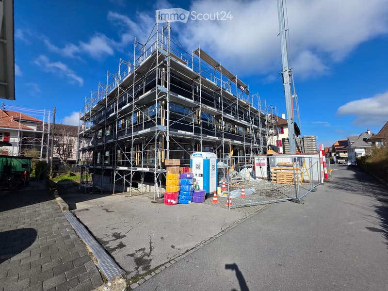 Multi-story building under construction with scaffolding, surrounded by temporary fencing, portable toilets, and stacked containers.