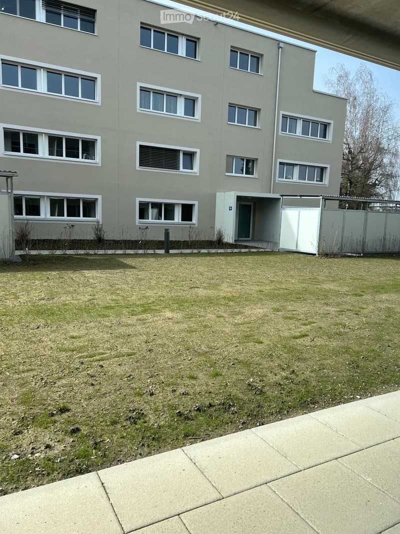 Modern multi-story building with light gray facade, multiple windows, and a small entrance area. The ground is covered with grass and concrete pavement.