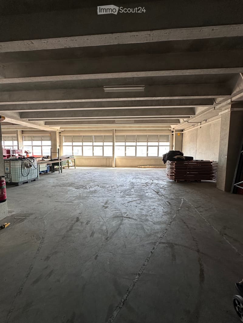 Empty industrial-style room with concrete floors and walls, large windows, and minimal furnishings including a few stacked items and a fire extinguisher.