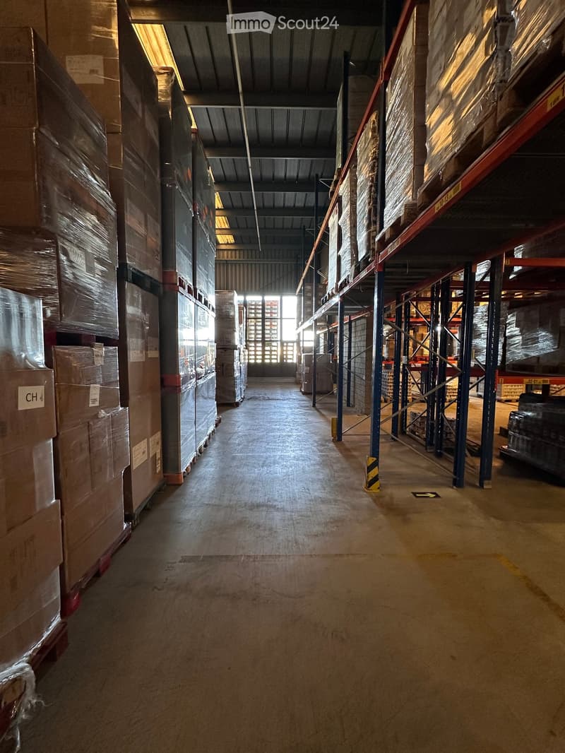 Warehouse corridor with high ceilings, metal shelving units, and stacked boxes on both sides. The floor is concrete, and there are industrial-style lighting fixtures on the ceiling.