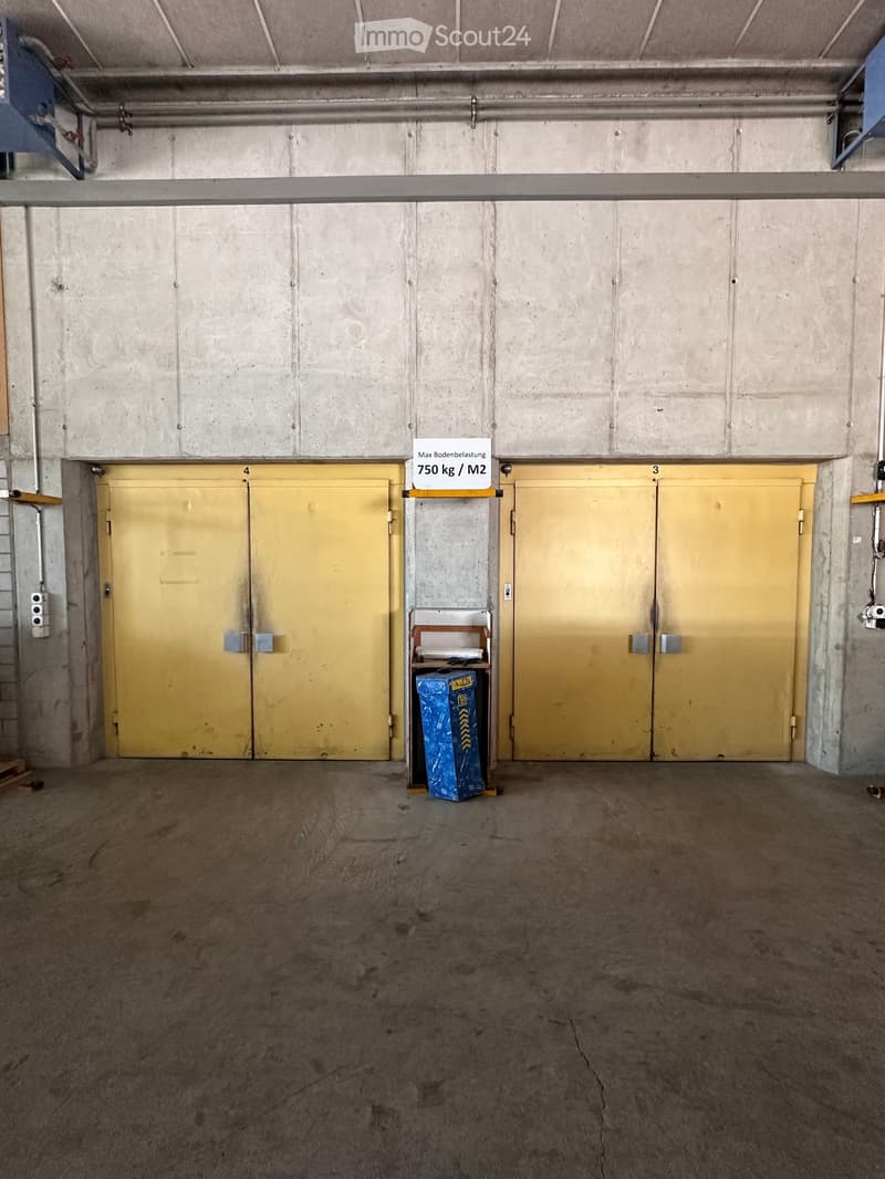 Empty underground parking space with two large yellow garage doors, concrete walls, and a small shelf with a blue recycling bin.