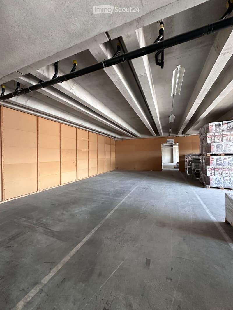 Empty concrete room with exposed pipes and lighting fixtures, partially covered walls, and storage crates in the corner.