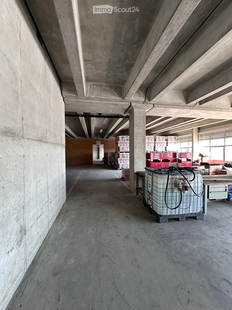 Concrete corridor with exposed beams and pillars, natural light from windows, storage containers and equipment visible in the background.