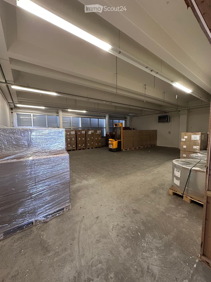 Empty warehouse space with concrete floor, fluorescent lighting, windows with blinds, and stacked boxes along the walls.
