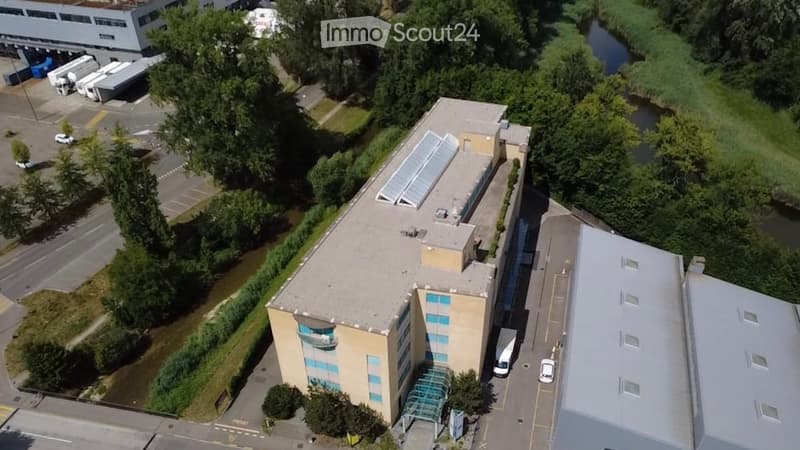 Aerial view of a modern multi-story building with a flat roof, surrounded by parking areas, greenery, and nearby industrial structures.