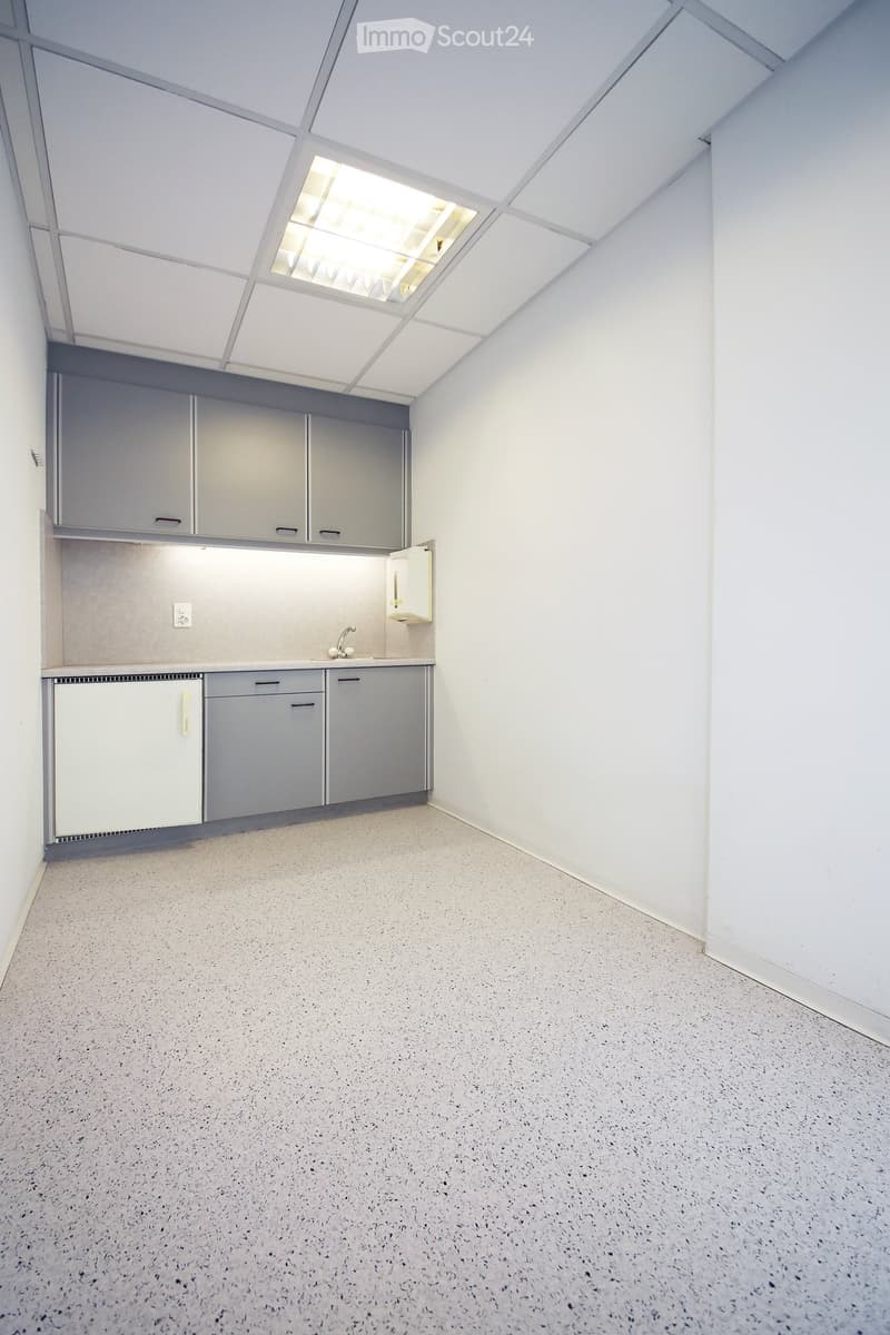 Empty room with grey cabinets, sink, and a small refrigerator. The ceiling has a skylight, and the floor is a speckled concrete surface.