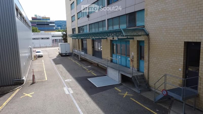 Multi-story building with a covered entrance, blue doors, and a paved parking area with marked spaces.