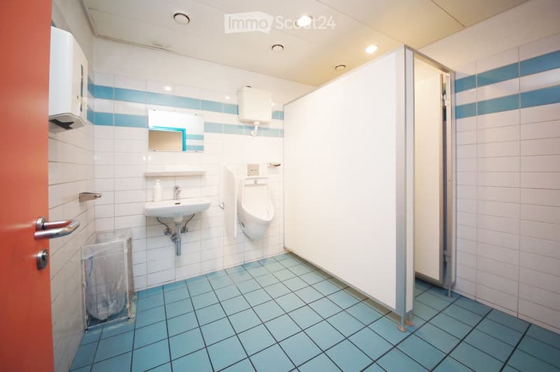 Clean public restroom with white and blue tiled walls, a sink, urinal, mirror, toilet paper dispenser, and a trash bin. The floor is covered with light blue tiles.