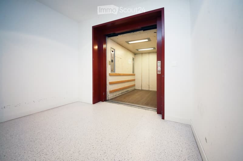 Empty room with white walls and a concrete floor, featuring an open doorway leading to another room with wooden flooring and a visible elevator shaft.