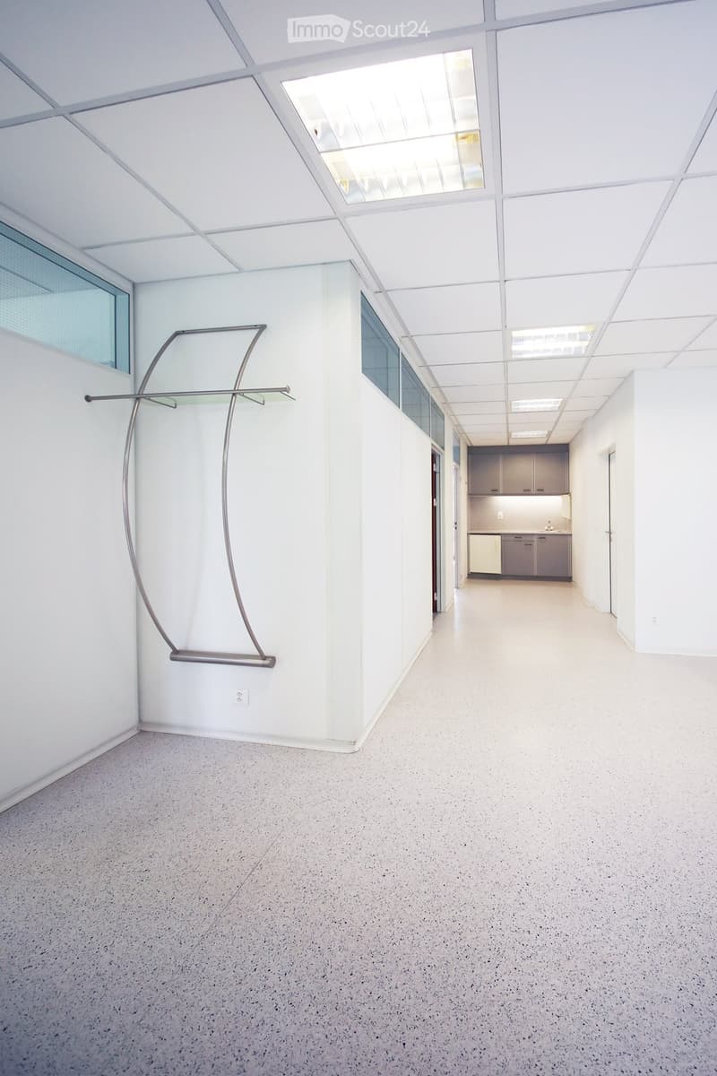 Open, modern corridor with white walls, a sleek floor lamp, and a small kitchenette area visible at the end.