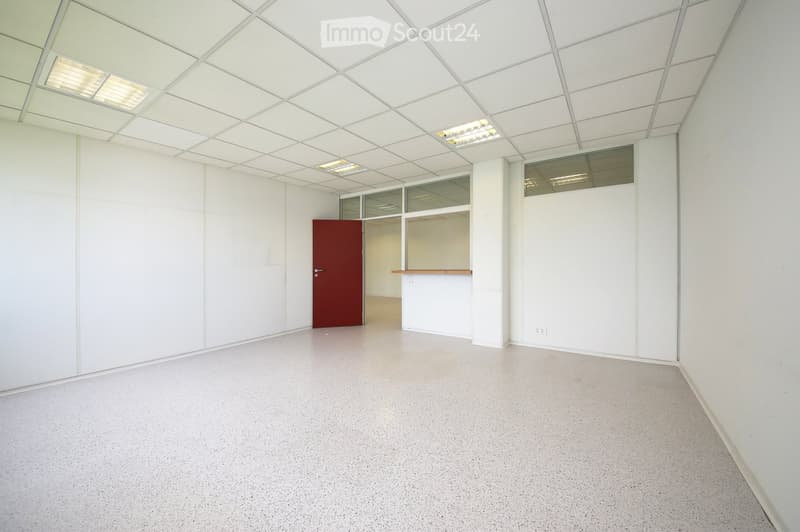 Empty room with white walls, grey carpeted floor, and a red door leading to another space. The ceiling has a grid pattern with integrated lighting.