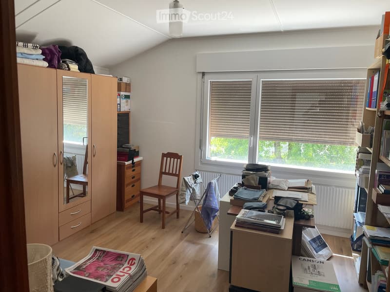 A well-lit attic room with wooden flooring, large windows, a slanted ceiling, and a skylight. The room features a wooden chair, a small table with a laptop, a cabinet with a mirror, a bookshelf, and various personal items including clothes, books, and a c