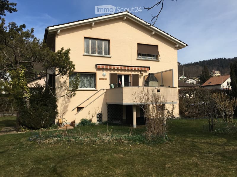 Two-story house with beige exterior, multiple windows, a covered balcony, and a small garden area.