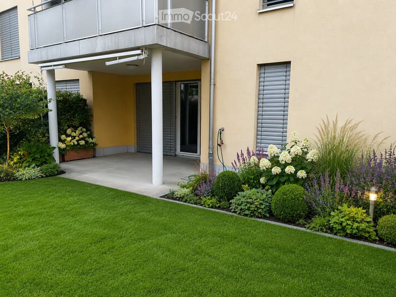 Well-maintained garden with green lawn, flower beds, shrubs, and a small pathway leading to a modern building entrance with a balcony and glass door.