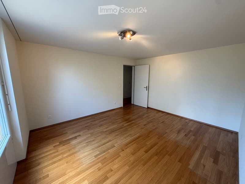 Empty room with light wooden flooring, white walls, and a ceiling light fixture. A white door is open to an adjacent space.