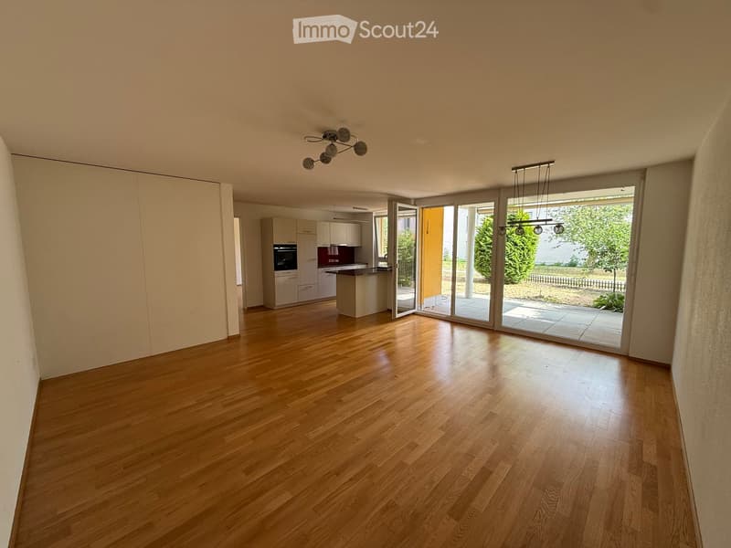Empty room with wooden flooring, large sliding glass doors leading to an outdoor area, and a modern kitchen visible in the background.