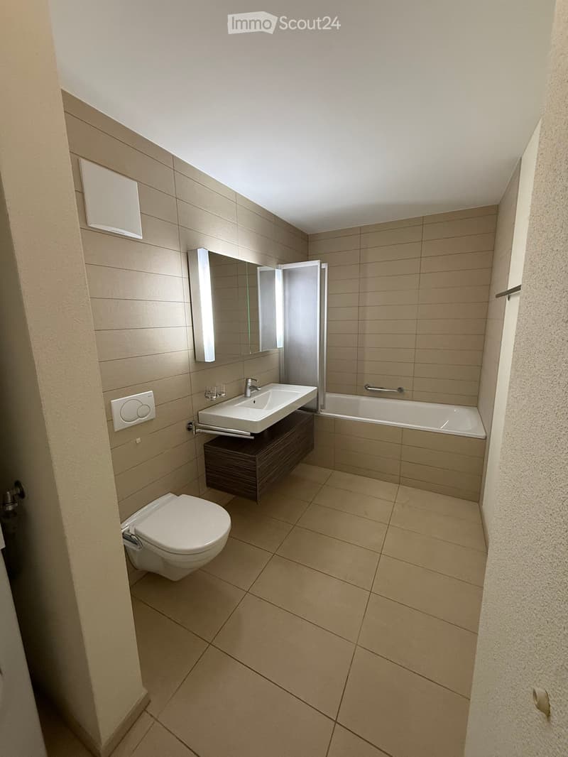 Modern bathroom with beige tiles, a white toilet, a sink with a mirrored cabinet, and a bathtub with a glass enclosure.