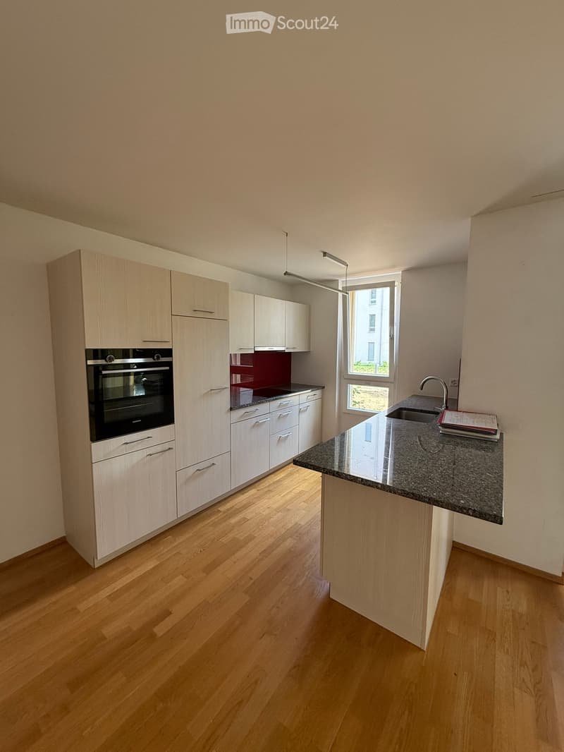 Modern kitchen with light wood cabinets, stainless steel oven, granite countertop, and a window overlooking a green area.