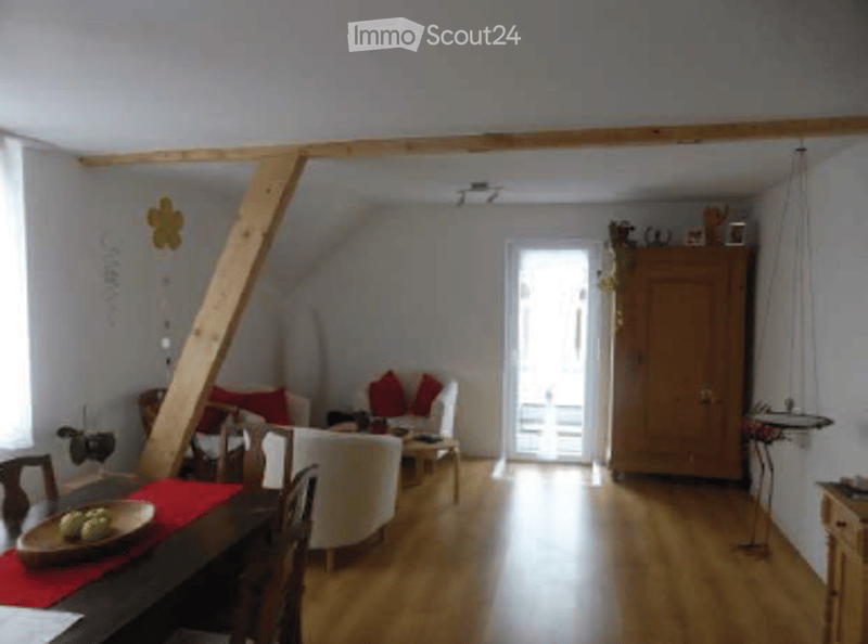 Bright, open-plan living and dining area with wooden flooring, white walls, and large windows. Features a sofa, dining table with chairs, and a wooden cupboard.